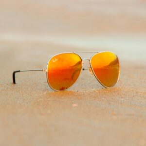 Close-up of aviator sunglasses on sand with orange reflection, evoking a warm summer vibe.